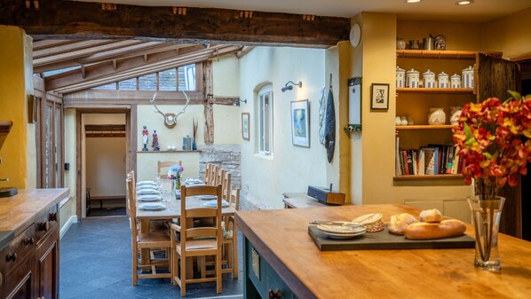 A rustic kitchen with wooden beams, a dining area with a wooden table and chairs, and a bookshelf with books and decorative items.