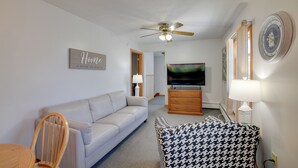 A living room with a sofa, a houndstooth armchair, a wooden chair, a television, a ceiling fan, and a clock on the wall. - Apt in Old Orchard Beach - Walk to Bay + Pier (Old Orchard Beach)