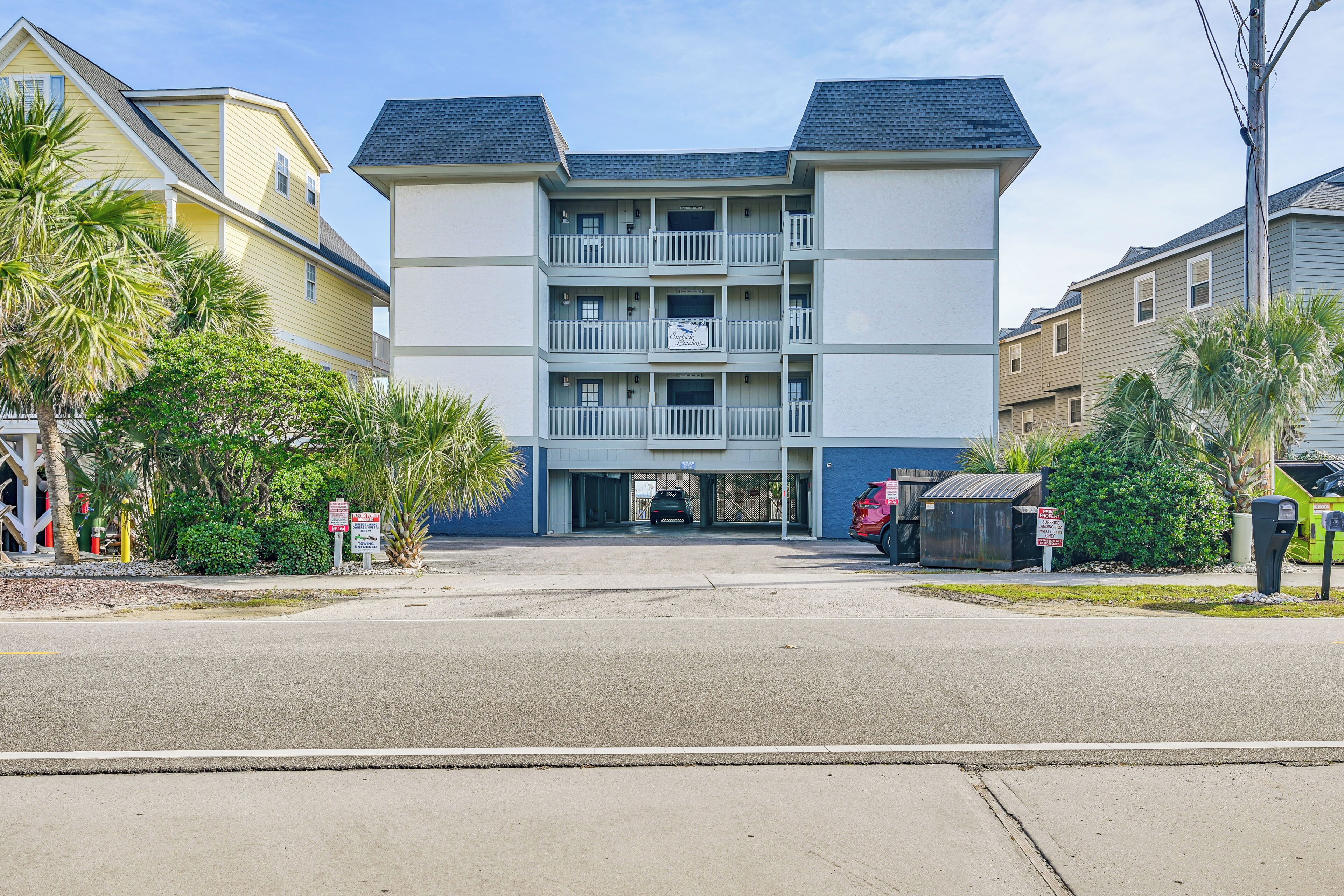A multi-story building with balconies, a parking lot, and surrounding vegetation.