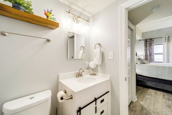 A bathroom with a white sink, mirror, and toilet. There is a wooden shelf above the sink with a potted plant. A towel hangs on a ring above the sink.