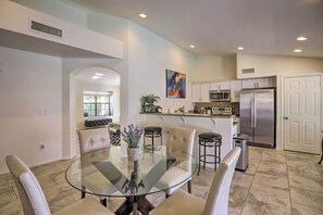 A modern kitchen with stainless steel appliances, a dining area with a glass table and cushioned chairs, and a living space with a sofa and a large window.
