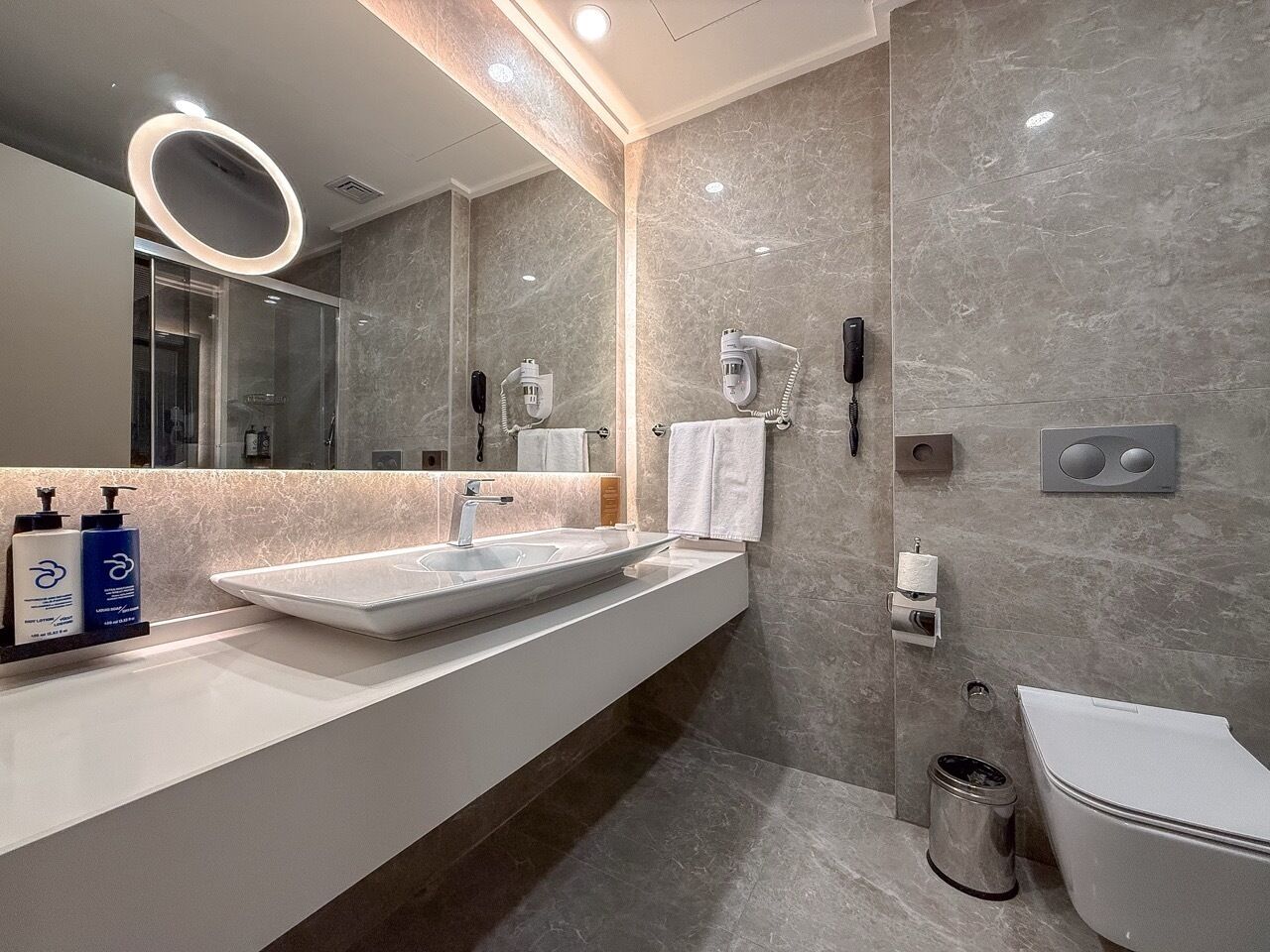 A modern bathroom with a large mirror, two sinks, a toilet, and a trash can.