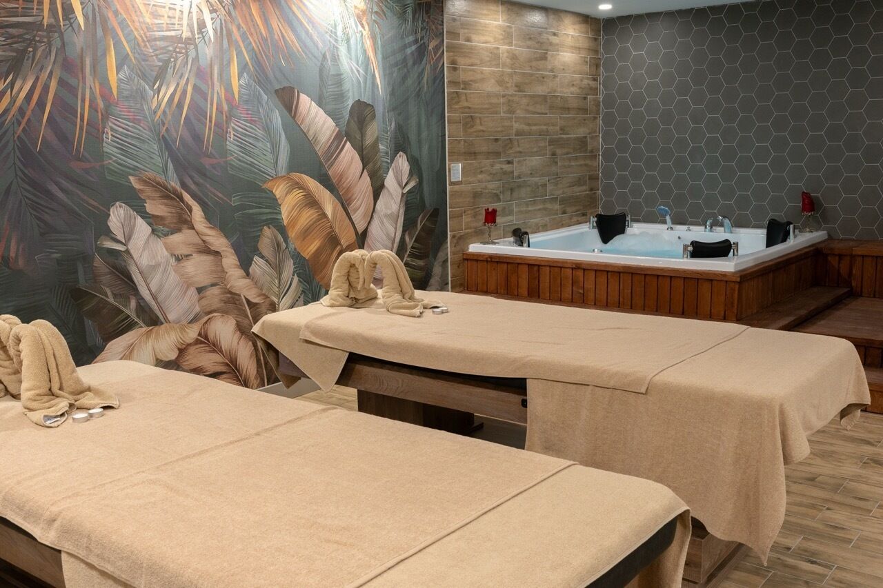A spa room with two treatment tables, a large mural of tropical leaves, a wooden wall, and a hexagonal tiled wall.