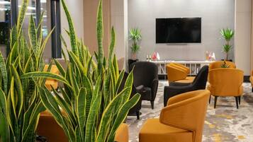 Un hall d’hôtel moderne, agrémenté d’une grande plante en pot, de sièges confortables, d’un téléviseur à écran plat et d’une table décorative ornée de drapeaux.