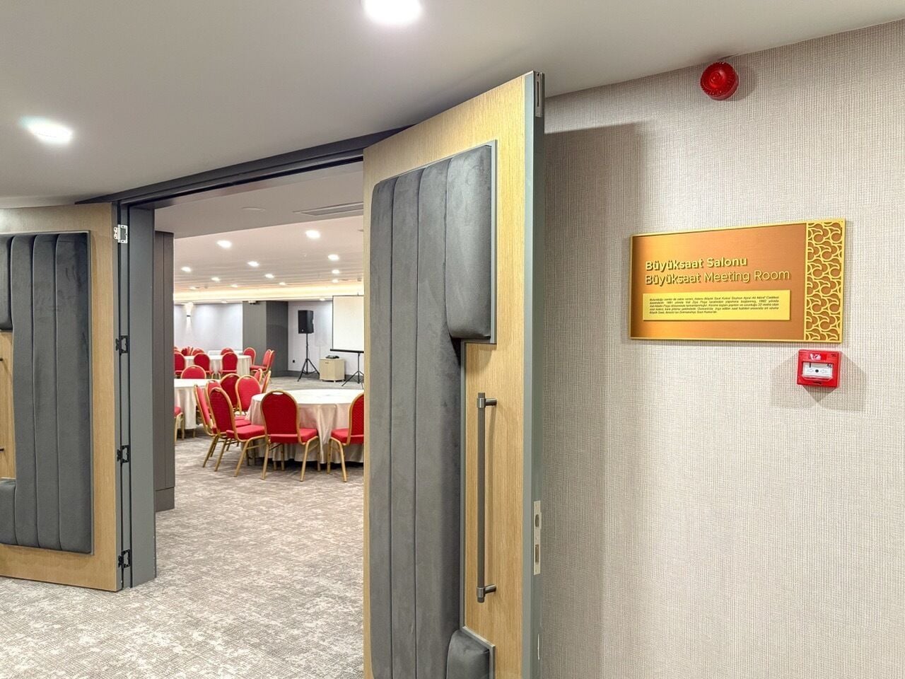 Una sala de reuniones con una puerta de madera, un letrero que la identifica como sala de reuniones y un botón rojo de emergencia en la pared.