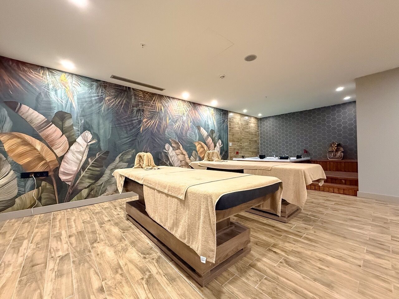 Ein Raum mit zwei Massagetischen, einer großen Wandmalerei mit tropischen Blättern und einem Empfangstresen.