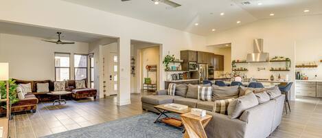 A spacious living room with a large sofa, a coffee table, and a dining area with chairs. There are ceiling fans, a kitchen with cabinets, and a large window.