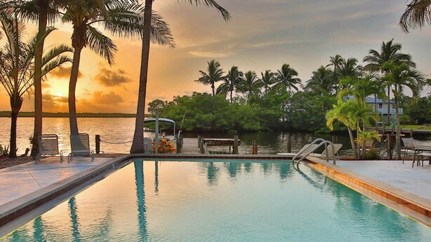 A swimming pool with lounge chairs and a dock, surrounded by palm trees and a view of the water. - Waterfront Matlacha Condo w/ Patio & Pool Access (Matlacha)