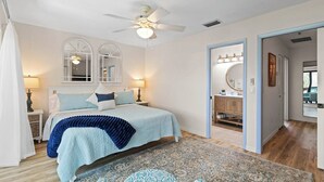 A bedroom with a bed, two bedside tables, a ceiling fan, a large window with a view, and a bathroom with a mirror and sink. - Waterfront Matlacha Condo w/ Patio & Pool Access (Matlacha)