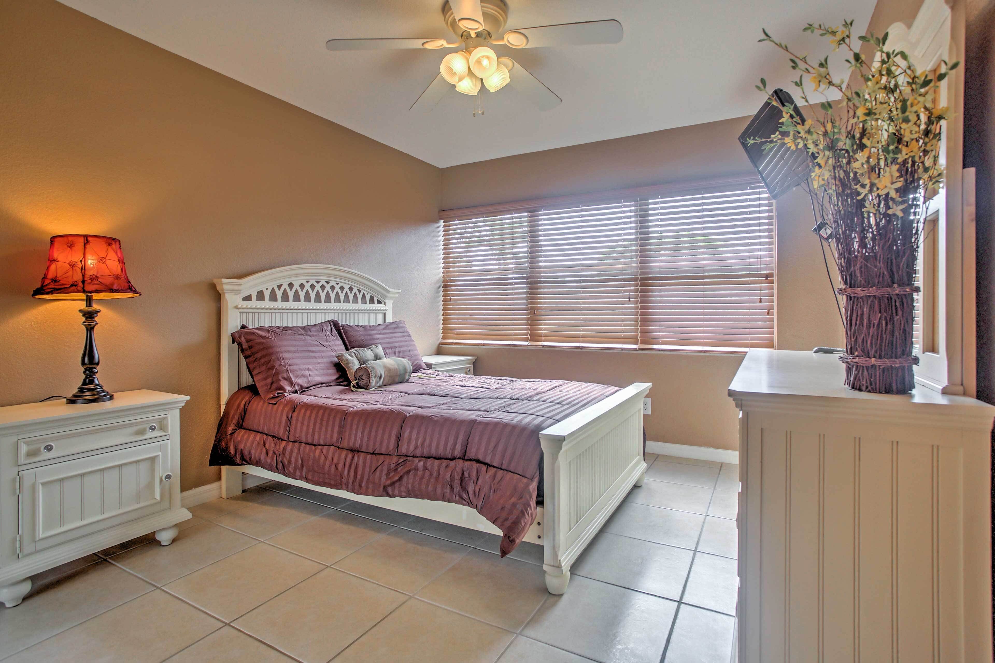 A bedroom with a bed, nightstand, lamp, ceiling fan, window with blinds, and a vase with flowers.