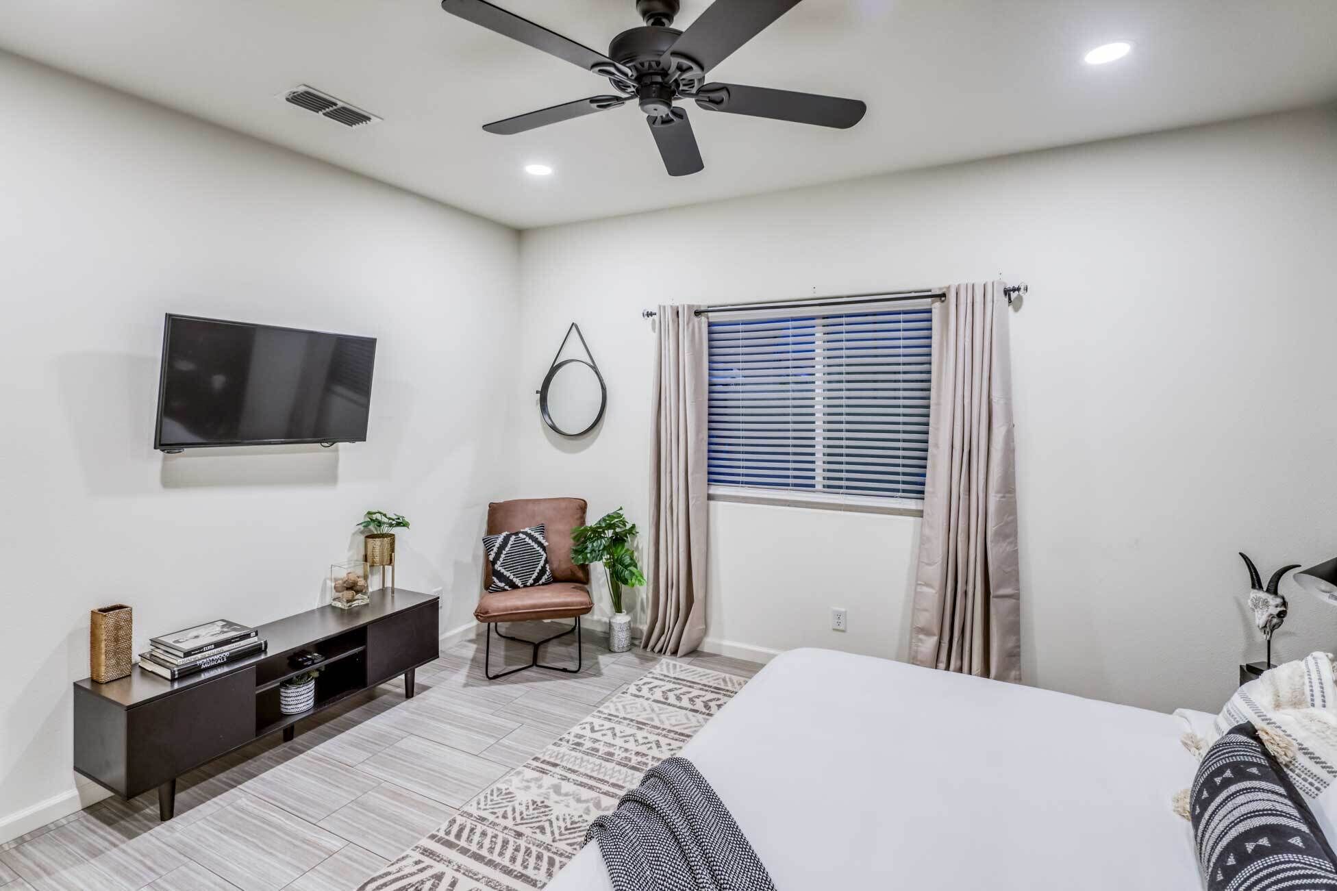 Un dormitorio con cama, televisión, ventilador de techo, ventana con cortinas, una silla y una pequeña mesa con objetos decorativos.