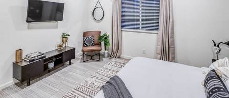 Un dormitorio con cama, televisión, ventilador de techo, ventana con cortinas, una silla y una pequeña mesa con objetos decorativos.