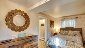 A bedroom with a wooden dresser, a circular mirror, a bed with patterned bedding, and a door leading to another room.