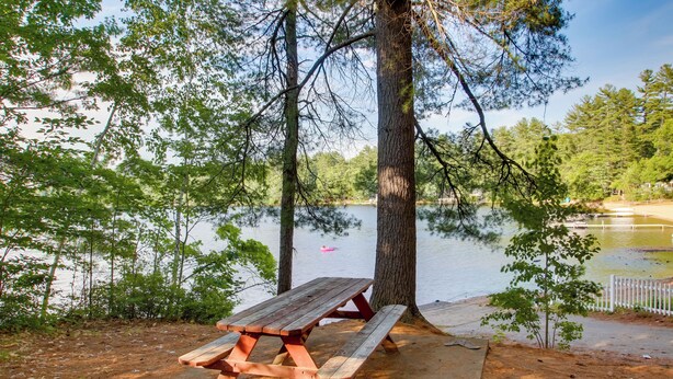 A picnic table by a lake with trees and a white fence in the background.