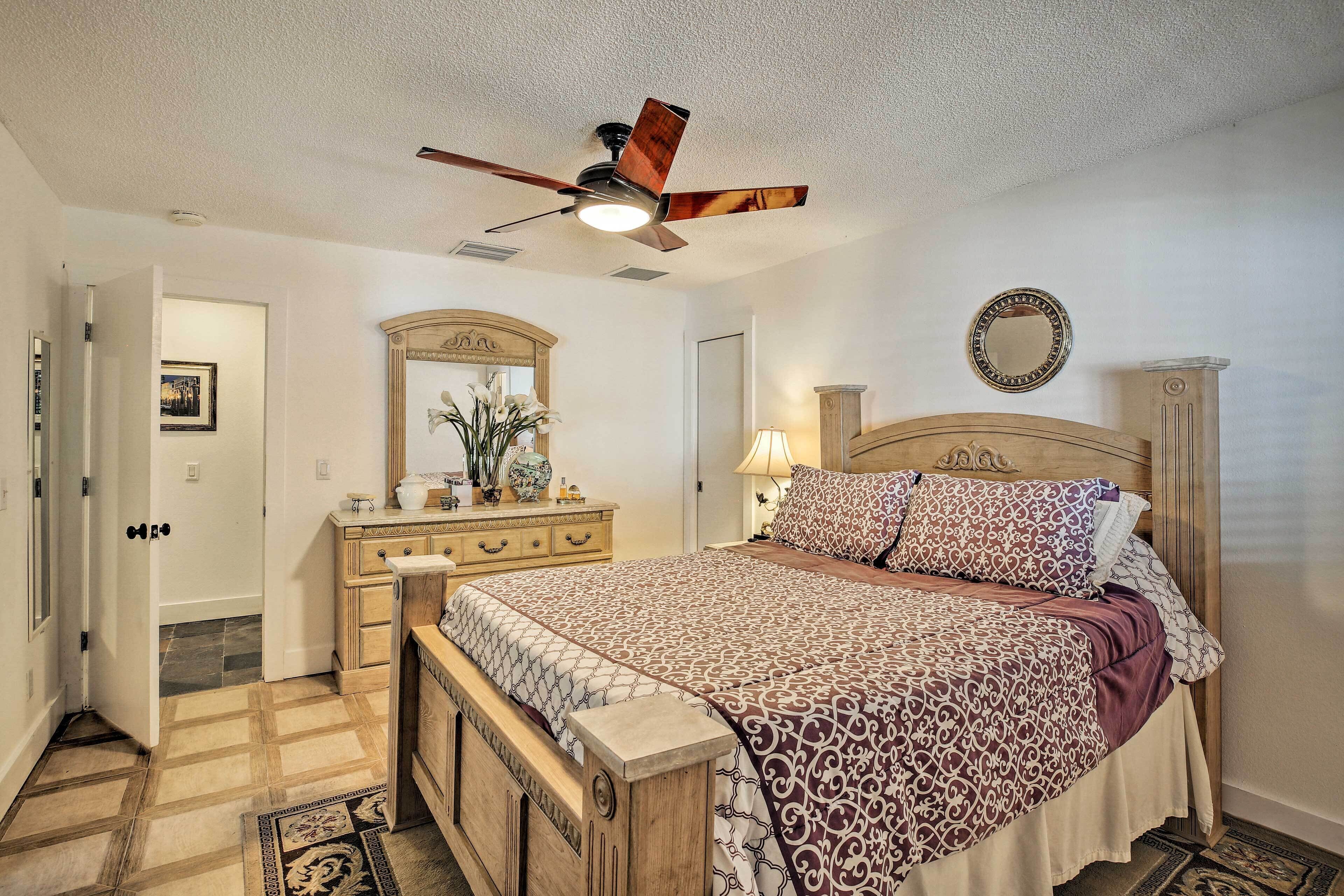A bedroom with a wooden bed, a ceiling fan, a dresser, a mirror, and a door leading to another room.
