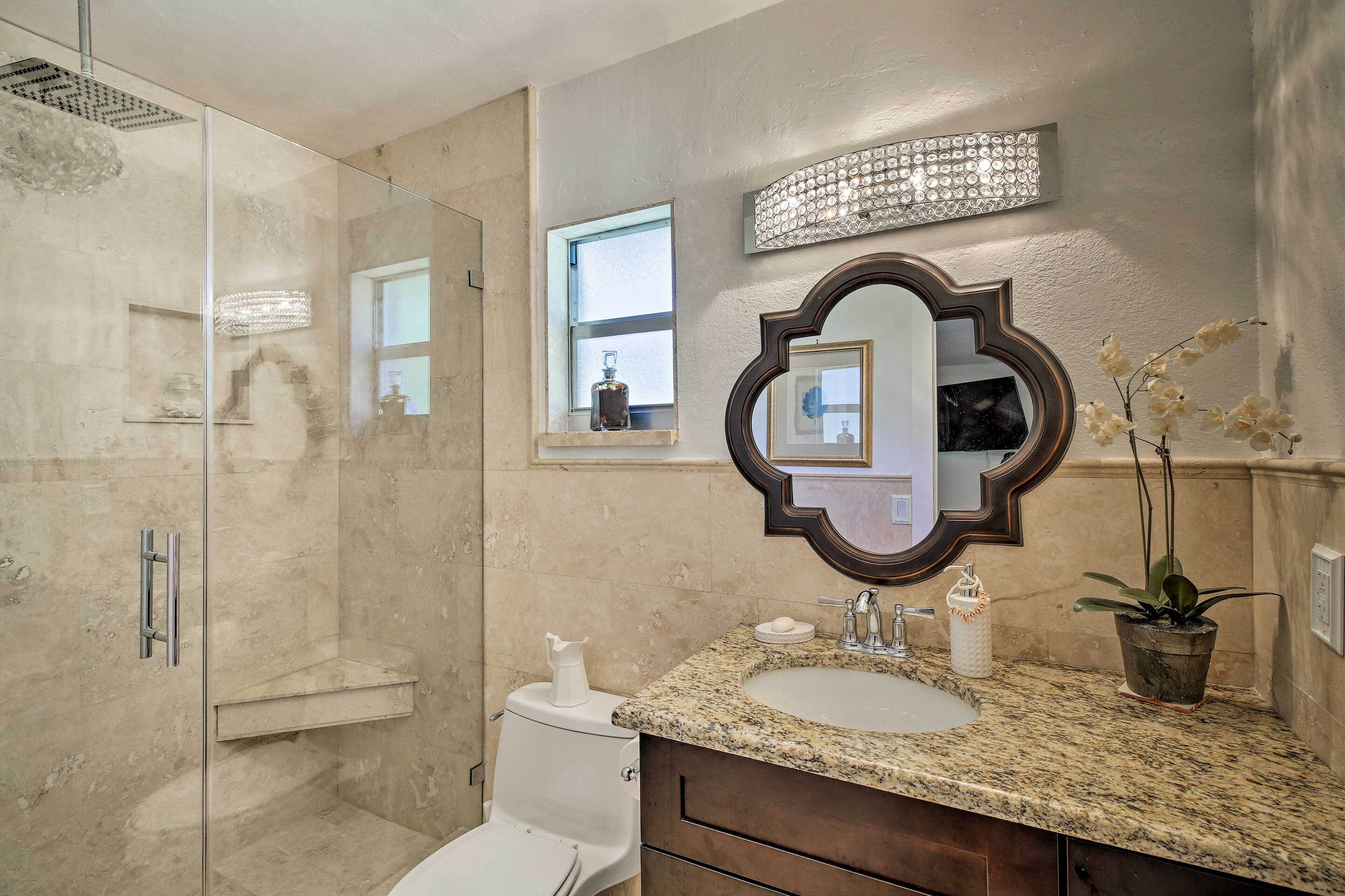 A bathroom with a glass-enclosed shower, a sink with a granite countertop, a mirror with a decorative frame, and a window with a view of the outside.