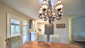 A dining area with a wooden table and four chairs, a chandelier, and a view of the kitchen and outdoor area through glass doors. - Brevard Outdoor Retreat at Connestee Falls (Brevard)