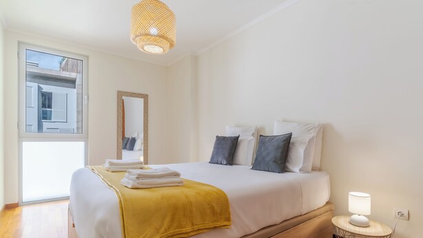A modern bedroom with a large bed, a bedside table with a lamp, a window with shutters, and a view of the outside. - Cozy Santa Cruz (Santa Cruz)