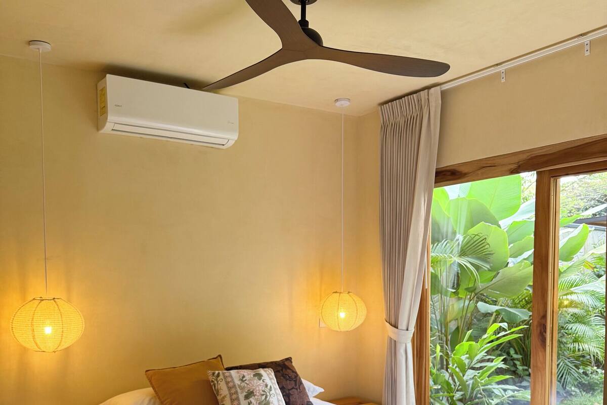 Ein Schlafzimmer mit Bett, Nachttischen, Deckenventilator, Klimaanlage und Blick auf Grün durch ein Fenster.