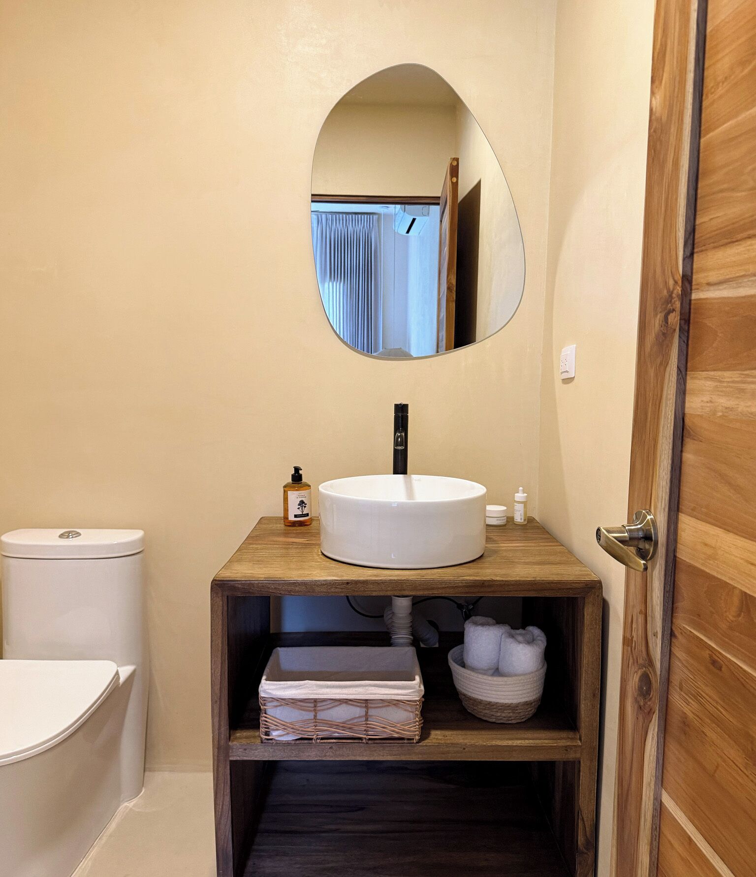 Un baño con tocador de madera, un lavamanos blanco, un espejo, un inodoro y un estante con toallas y artículos de aseo.