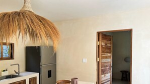 Une cuisine moderne avec un réfrigérateur en acier inoxydable, un meuble en bois, un plan de travail blanc, un luminaire en fibres tressées et une porte en bois donnant sur une autre pièce.