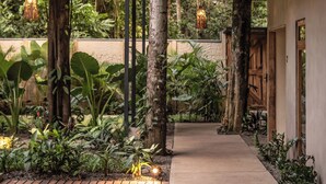 Un espace extérieur abrité, doté de poutres en bois et de luminaires suspendus, entouré d’une végétation luxuriante et bordé d’un chemin en béton.