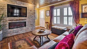 A living room with a stone fireplace, a flat-screen TV, and a large window with curtains. - Lovely Cottage w/ Sauna & Fire Pit Near Asheville! (Swannanoa)