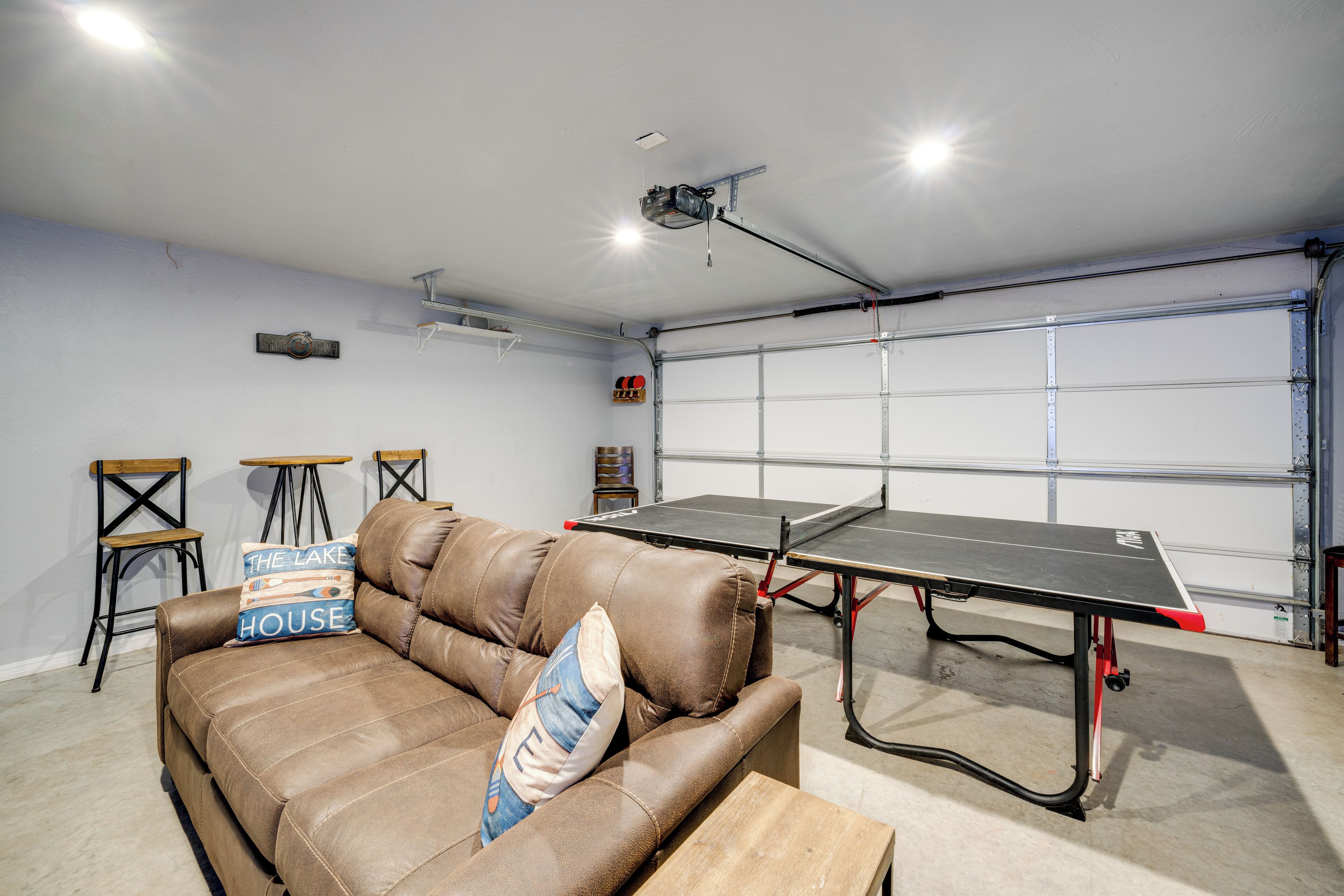 A room with a brown leather couch, a ping pong table, and a garage door in the background.