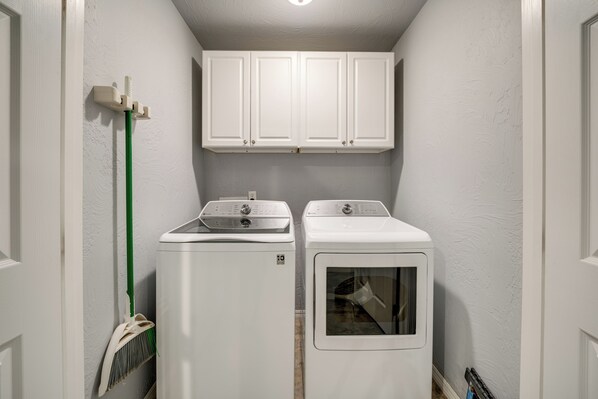 A laundry room with two front-loading washers and dryers, white cabinets above, and a broom leaning against the wall. - Walk to Lake Texoma! Modern Retreat w/ Game Room (Kingston)