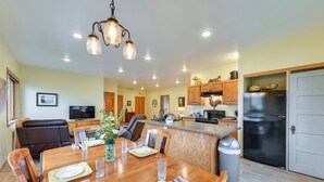 A dining area with a wooden table set for four, a kitchen with a refrigerator and cabinets, and a TV in the background. - Spacious Home 10 Minutes to Ten Sleep Canyon! (Ten Sleep)