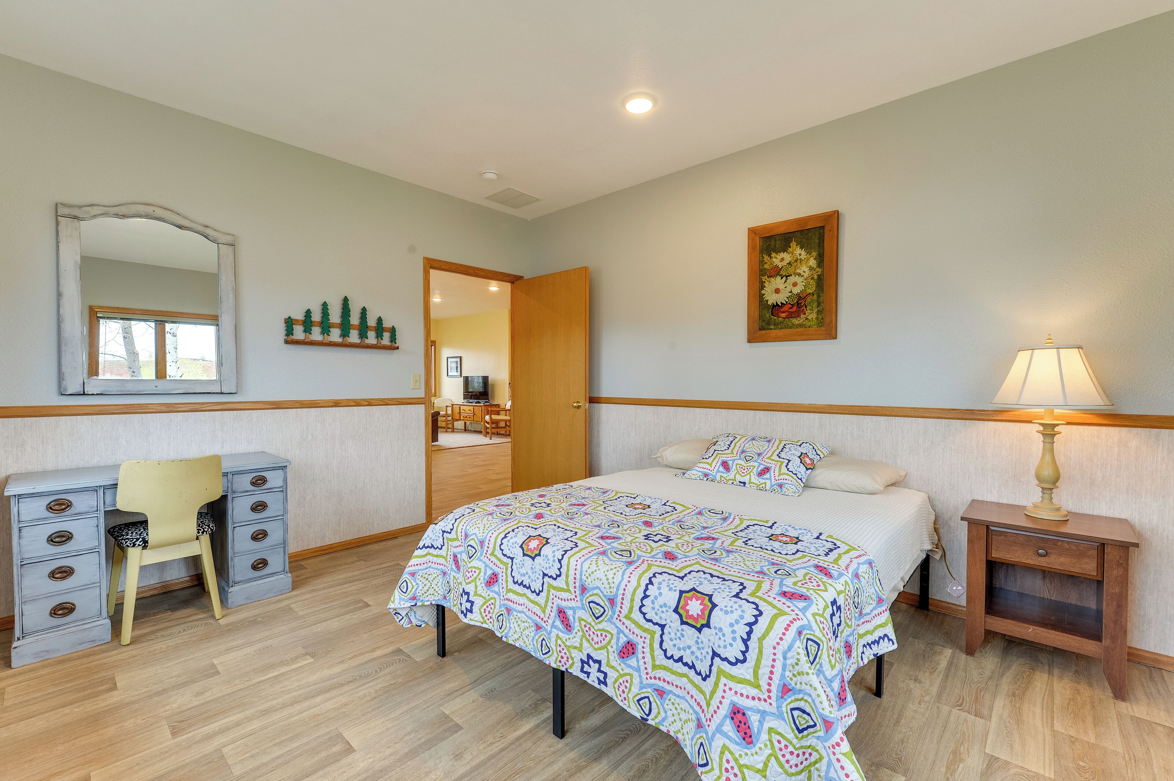 A bedroom with a bed, a desk with drawers, a chair, a bedside table with a lamp, and a mirror on the wall.