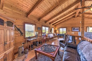 Interior de una cabaña de troncos con paredes de madera, techo alto, chimenea, sofá de cuero y mesa de comedor.