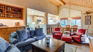 A spacious living room with a dark wooden coffee table, blue sofa, and red leather chairs. A kitchen is visible in the background with wooden cabinets and a microwave.