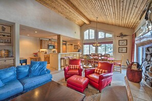 Ein gemütliches Wohnzimmer mit einem blauen Sofa, roten Ledersesseln und einem Couchtisch aus Holz. Im Hintergrund ist eine Küche erkennbar, mit Holzschränken und einem Essbereich mit kariertem ro-weißen Tischlein.