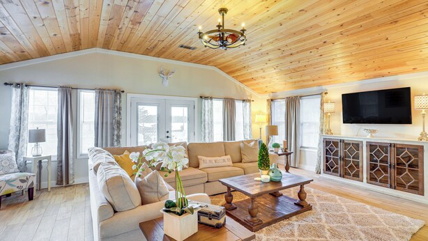 A spacious living room with a high wooden ceiling, a large sofa, a coffee table, and a television mounted on the wall. - Lake Sinclair: Since Day 1 Family Lake House (Milledgeville)
