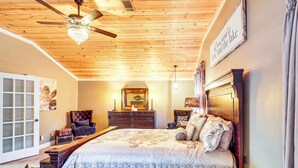 A bedroom with a wooden ceiling, a bed with a floral bedspread, a dresser, a mirror, and a chandelier. - Lake Sinclair: Since Day 1 Family Lake House (Milledgeville)