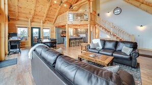 A spacious living area with a high ceiling, wooden beams, a staircase, a dining area, and a kitchen. - Large Deck & Shared Dock: Cabin on Kentucky Lake! (Gilbertsville)