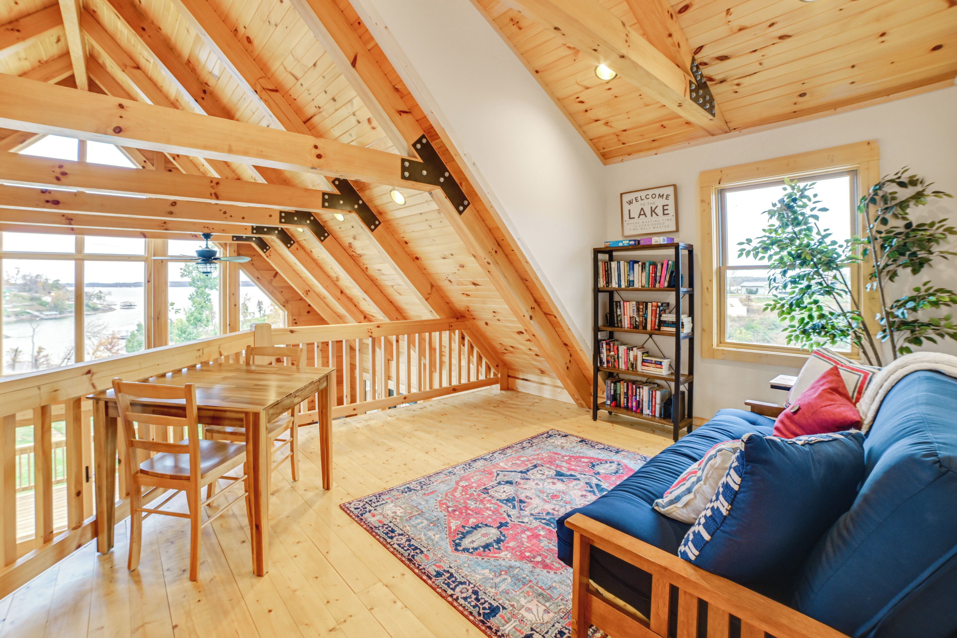 A cozy loft-style room with a wooden ceiling, a dining table with chairs, a blue sofa, a bookshelf, and a large window.