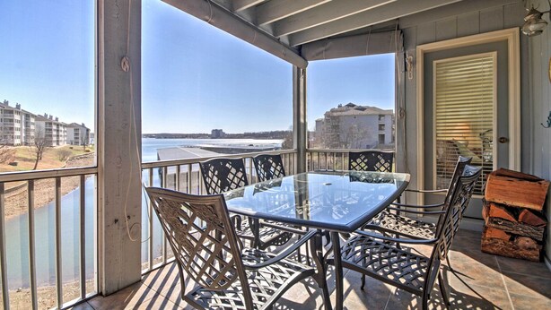 Een overdekt balkon met een glazen tafel en stoelen, met uitzicht op een waterpartij en gebouwen.