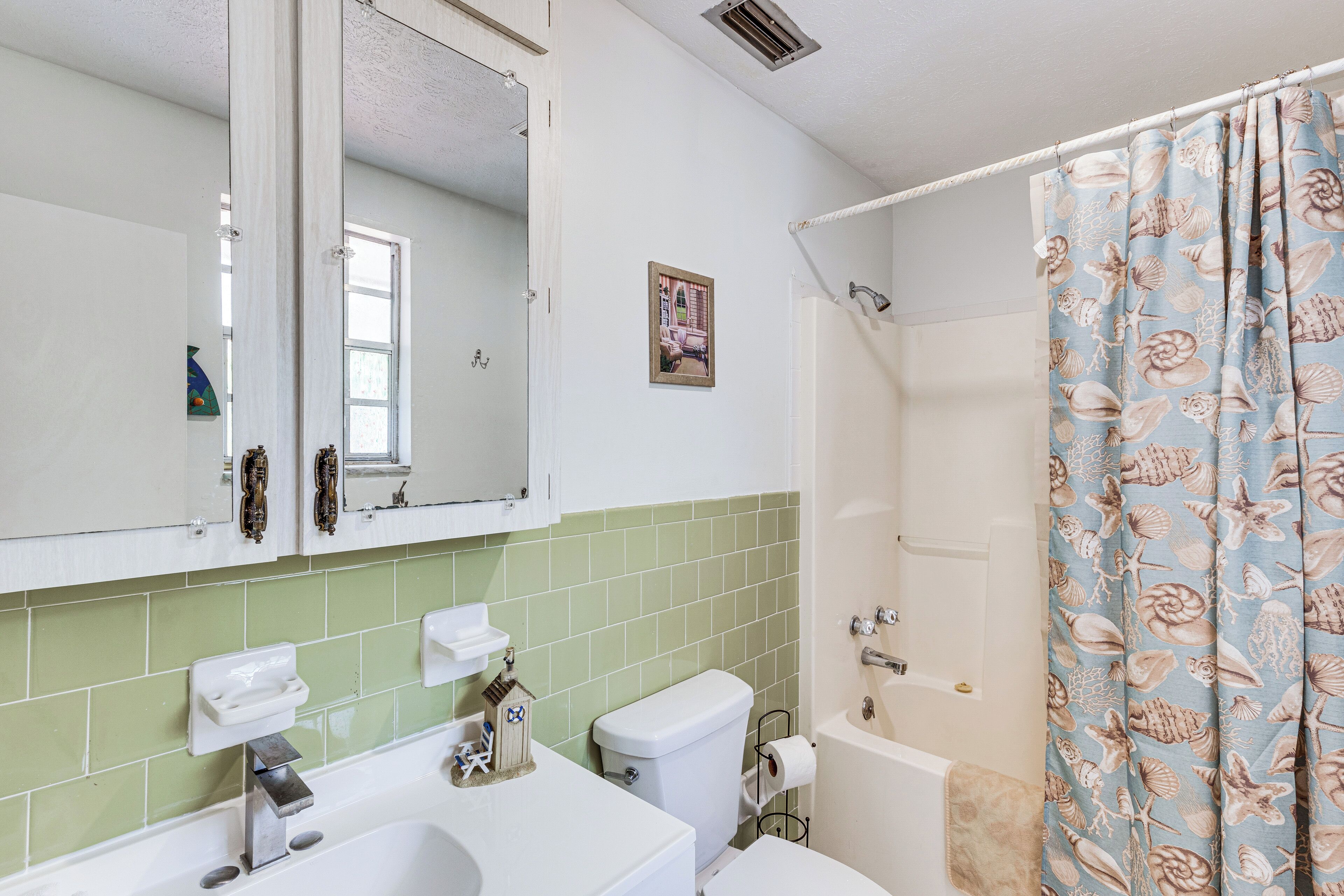 Un baño con lavamanos blanco, inodoro, bañera y cortina de ducha con diseño de conchas marinas.