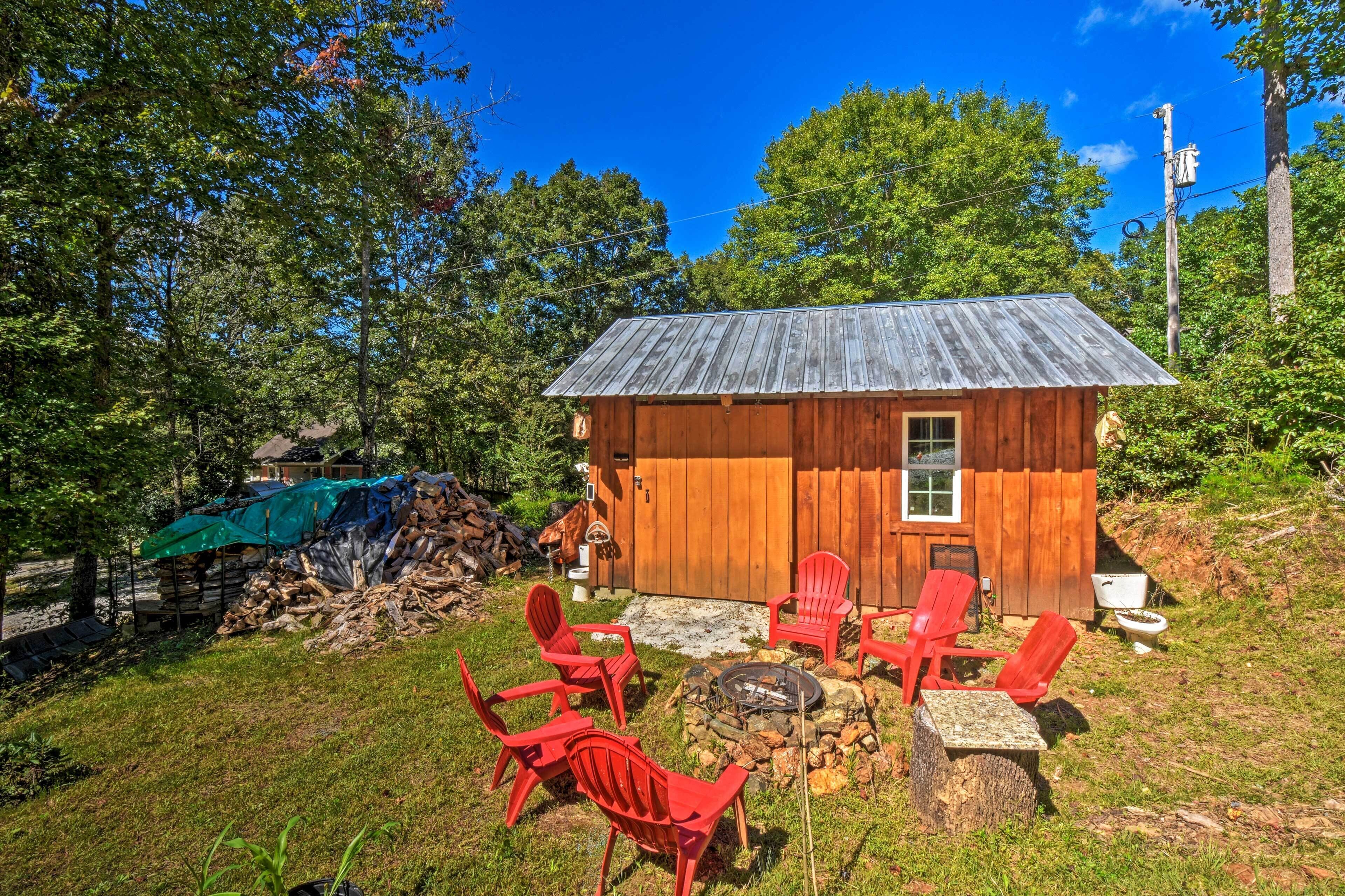 A wooden cabin with a metal roof, surrounded by a grassy area with red Adirondack chairs and a stone fire pit.