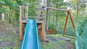 A wooden playground with a slide and swings set in a wooded area. - Georgia Home w/ 2 Fire Pits & Creek Access! (Young Harris)
