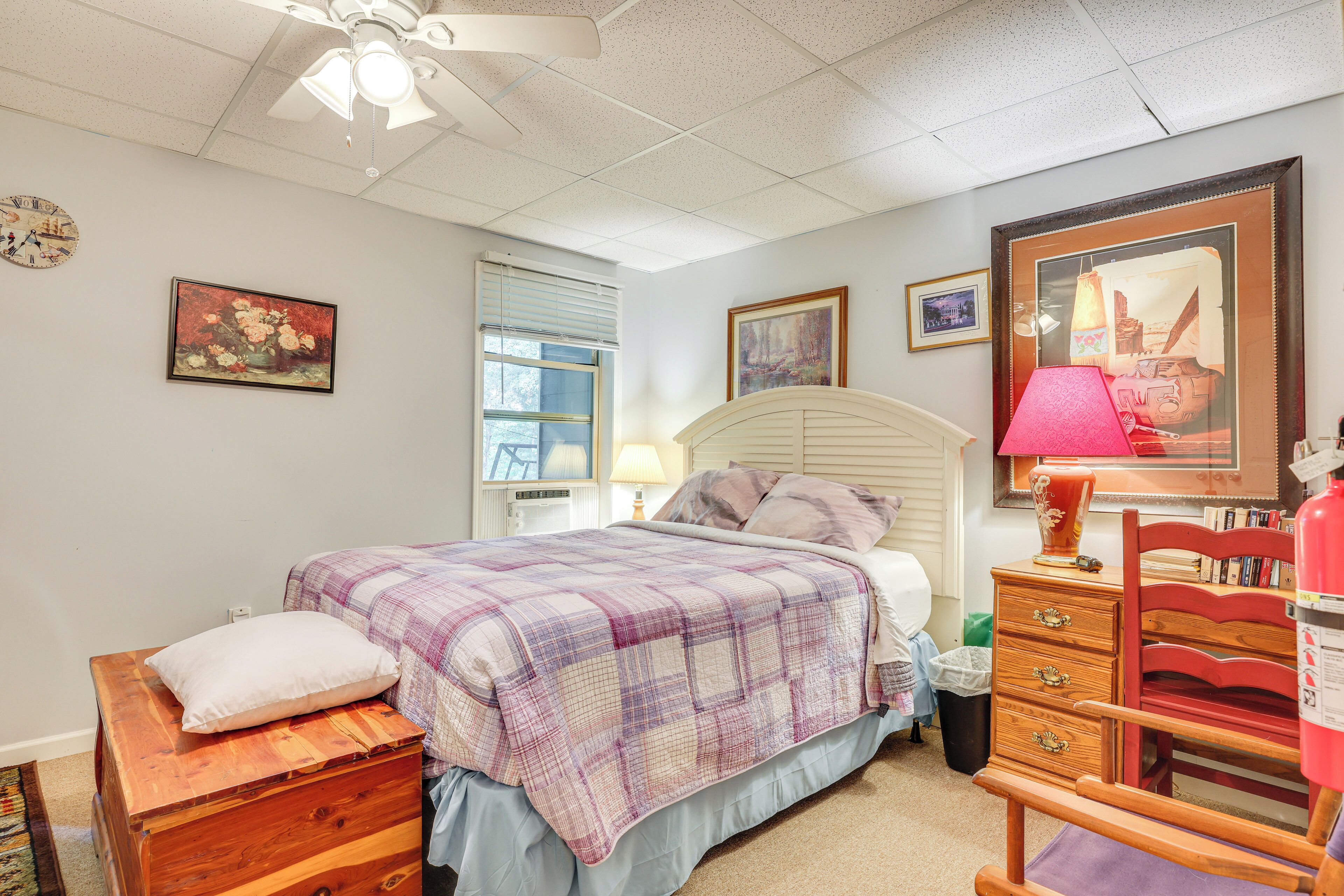 A bedroom with a bed, dresser, and a chair. There is a window with blinds, a ceiling fan, and a clock on the wall.