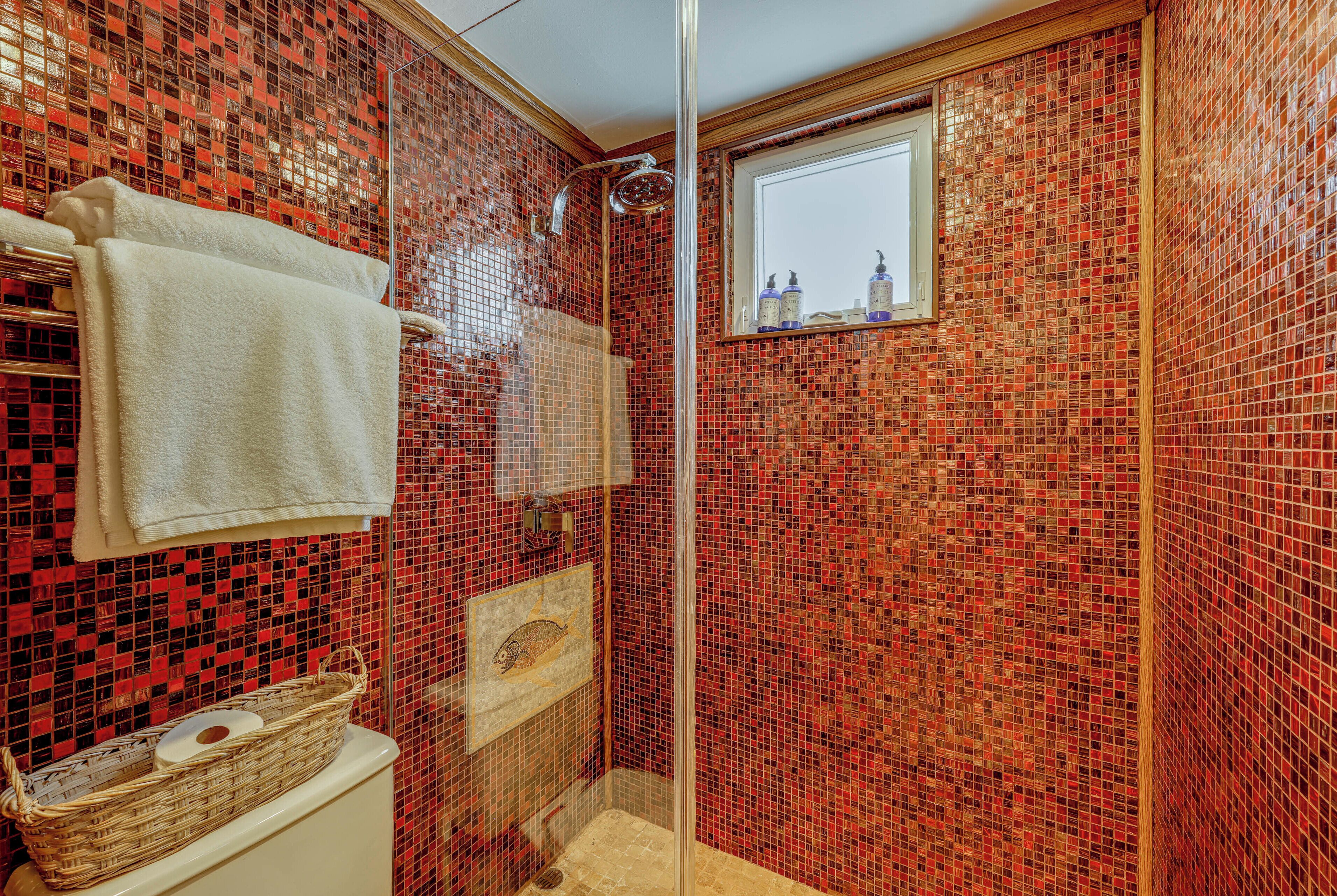 Un baño con mosaicos rojos, una cabina de ducha de vidrio, un lavabo blanco con una cesta y una ventana sobre el espejo.