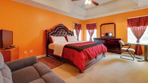 Een slaapkamer met een oranje wand, een bed met een rood en zwart dekbedovertrek, een houten hoofdeinde, een plafondventilator, een bureau met een spiegel, een stoel en een televisietoestel op een dressoir.