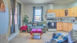 A compact living space with a sofa, a table, a kitchen area with wooden cabinets, and a window with curtains. - Rejuvenating Retreat w/ Ideal Eloy Location! (Eloy)