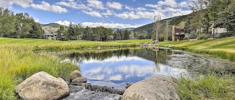 A serene lakeside view with a small stream, surrounded by lush greenery and mountains in the background.