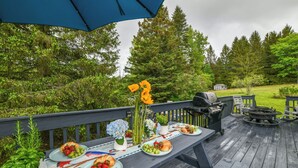 Un área de comedor al aire libre con terraza de madera, una parrilla y una mesa dispuesta con platos de comida y flores.