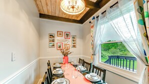 Comedor con mesa dispuesta para seis, candelabro, cuadros en la pared y ventana con vista a follaje.