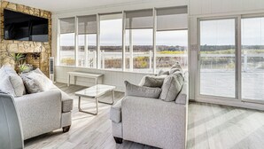 Ein modernes Wohnzimmer mit Kamin aus Naturstein, zwei grauen Sofas, einem Couchtisch und großen Fenstern mit Blick auf ein Gewässer und die Ferne.